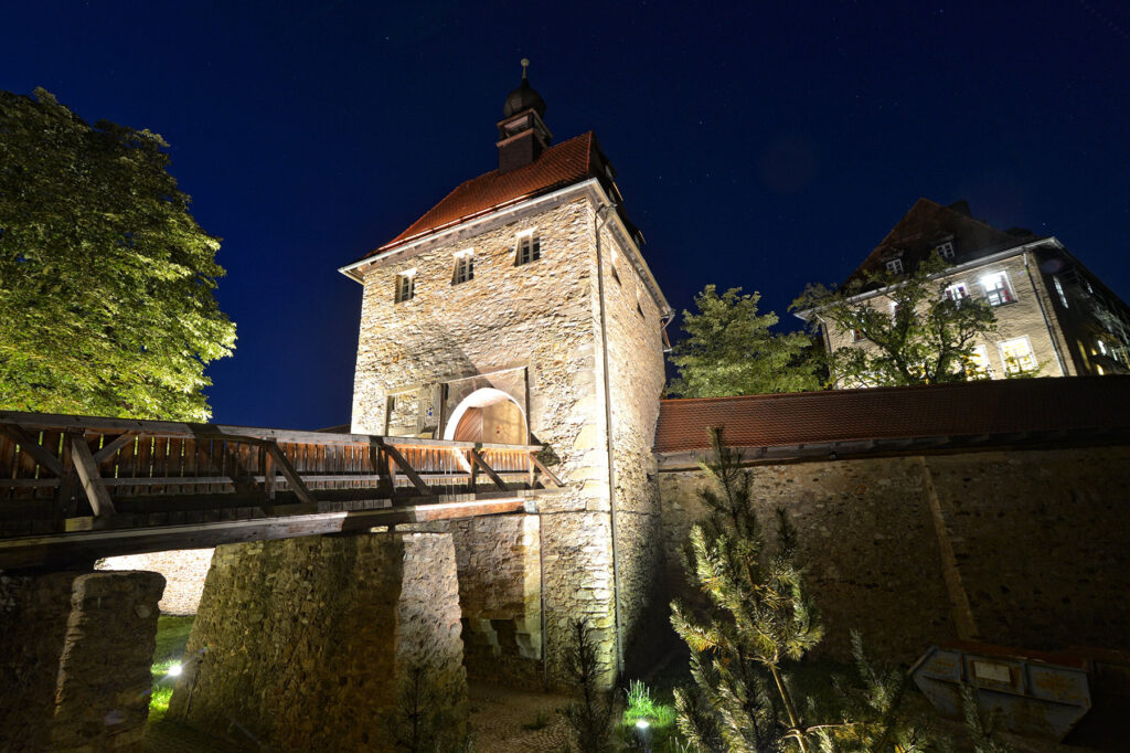 Burg Hohenberg an der Eger in dunkler Nacht, beleuchtete Steinmauer und Türmchen über einer Holzbrücke, Blick ins Egertal, historischer Ortskern