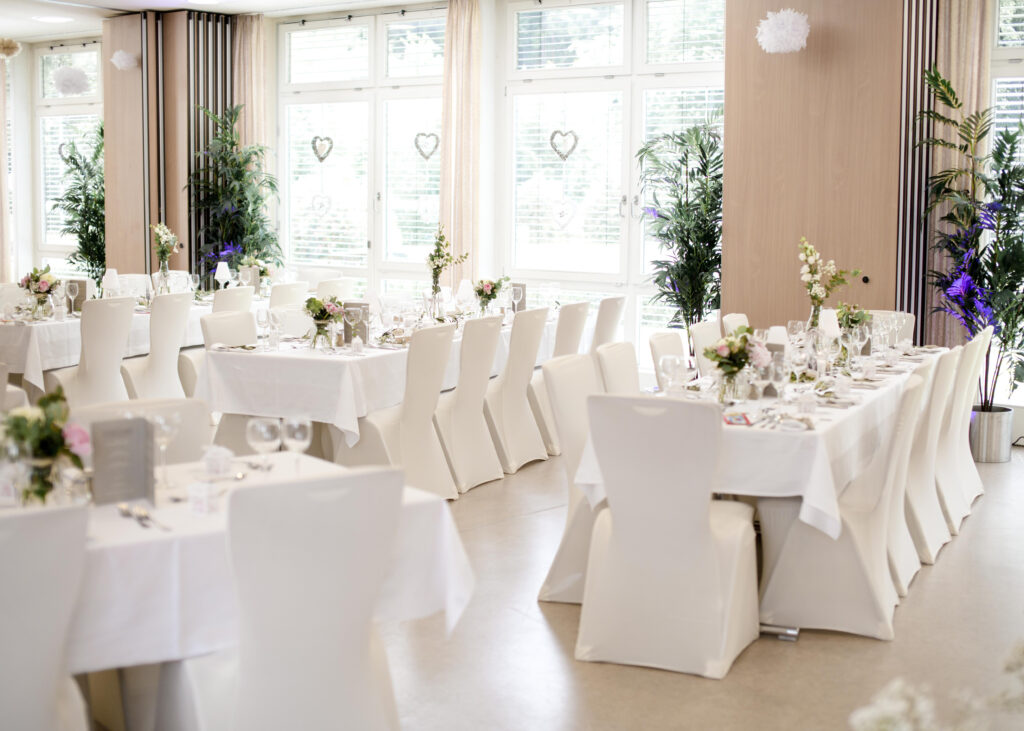 Weiß eingedeckte Tische mit Blumengestecken und Herz-Deko im Festsaal, große Fenster, grüne Pflanzen; Ambiente im Haus Silberbach