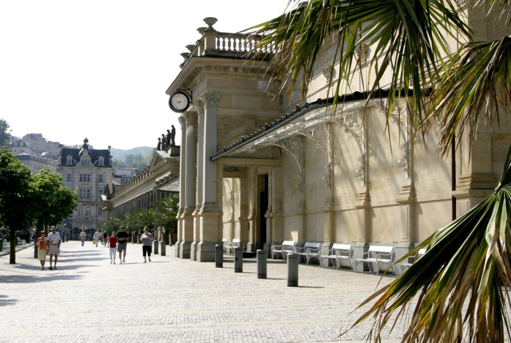 Karlovy Vary Kurpromenade mit hellen Sandsteinfassaden, Arkaden, Uhrturm, Palmen und Besucher auf einem sonnigen öffentlichen Platz – eine elegante historische Baukulisse.