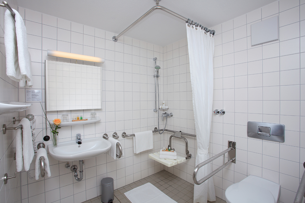 Badezimmer mit weißen Keramikwaschbecken, Spiegel, Dusche, Vorhang und Handläufen, rollstuhlgerechter Zugang im Hotel Haus Silberbach, Appartements. Rollibad.