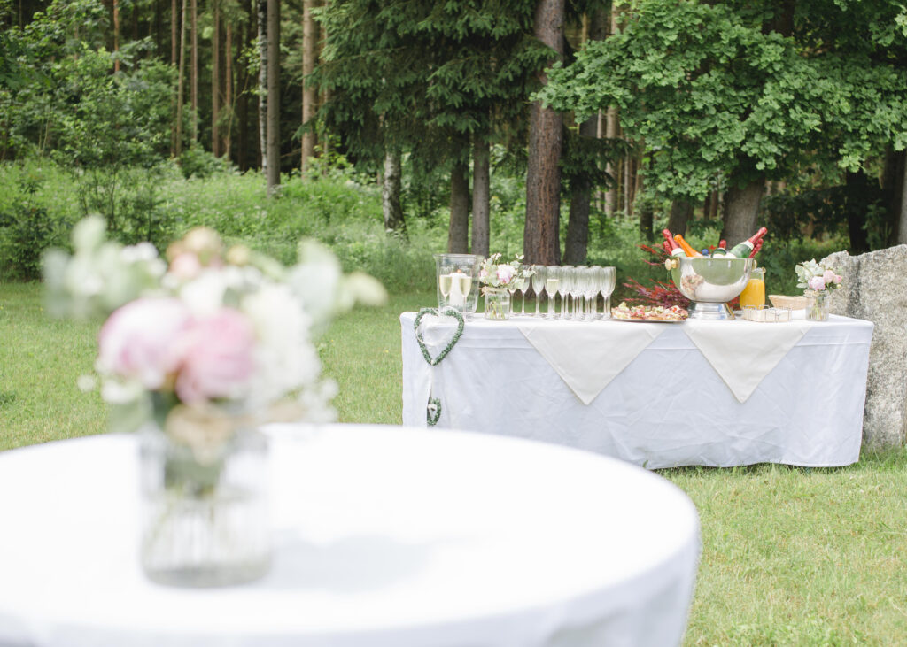 Garten-Sektempfang vor Haus Silberbach: ein weiß eingedeckter Tisch mit Sektgläsern, Bowle- oder Saftkaraffe, Gratulieren-Girlande und eine Vogelperspektive auf grünen Wald im Hintergrund