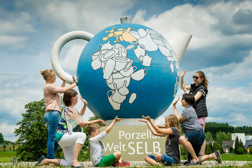 Große blaue Porzellankanne-Skulptur der Porzellanwelt Selb, umgeben von spielenden Besuchern; Landmarke am Naturpark Fichtelgebirge nahe Haus Silberbach.