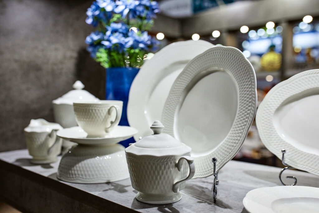 Weiße Porzellan-Geschirrset mit Tassen, Deckel und Platten in einem Ausstellungsdisplay, blaues Blumenarrangement im Hintergrund; Marken Rosenthal und Villeroy & Boch sichtbar.