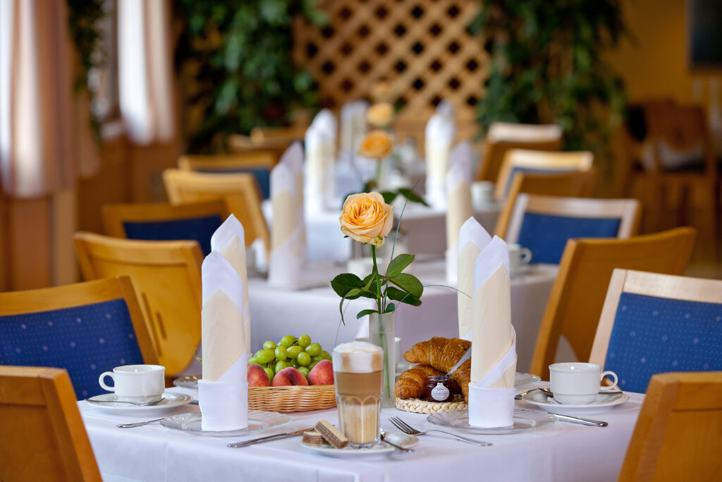 Tisch im Hotel Haus Silberbach mit weißem Tischtuch, orange Rose in Vase, Fruchtkorb, Croissant und Cappuccino; warme Holzstühle