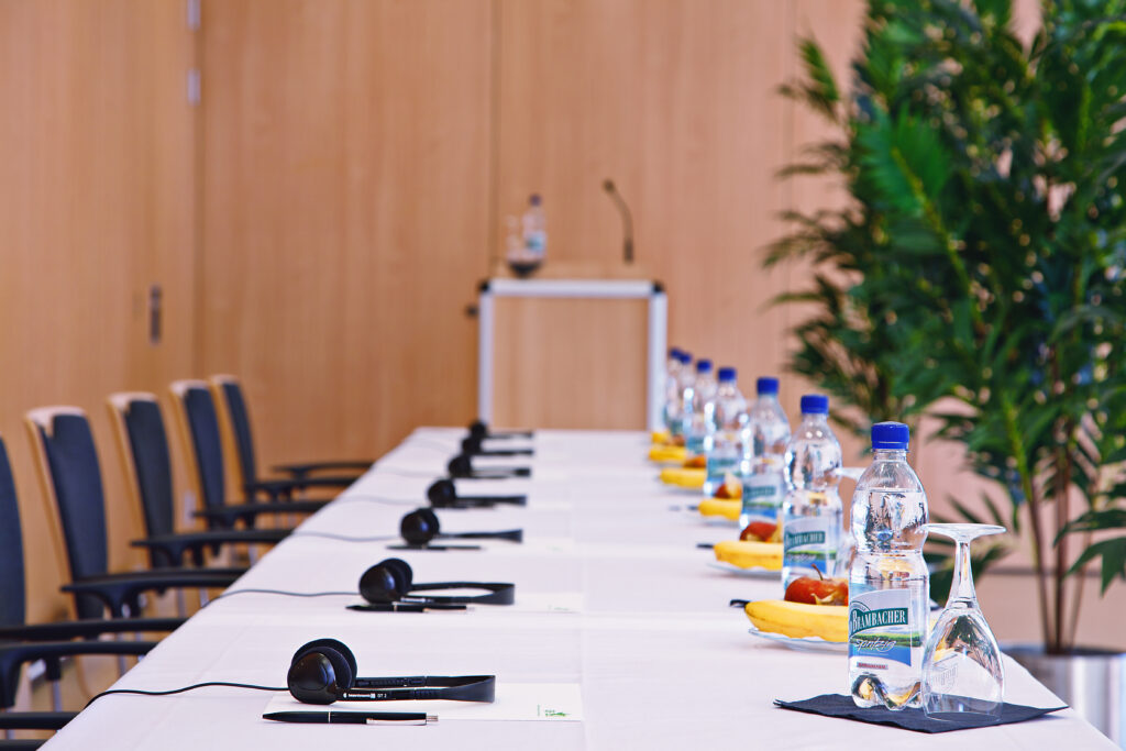 Stillleben eines Konferenzraums: Dolmetscheranlage, Headsets, Mikrofon, Wasserflaschen und Notizmaterial auf einem langen Tagungstisch; Holzverkleidung im Hintergrund und grüne Pflanzen im Raum