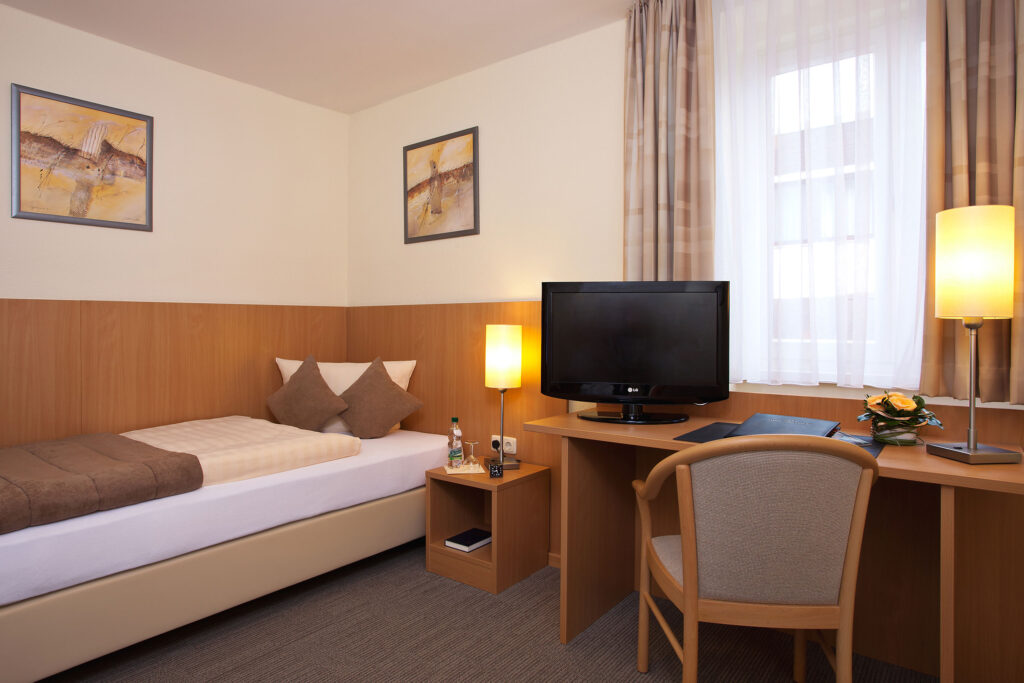 Beige-braunes Hotelzimmer im Haus Silberbach in Selb mit Bett, Kissen, Flachbild-TV, Schreibtisch, Lampe, Vorhängen und dekorativen Kunstwerken