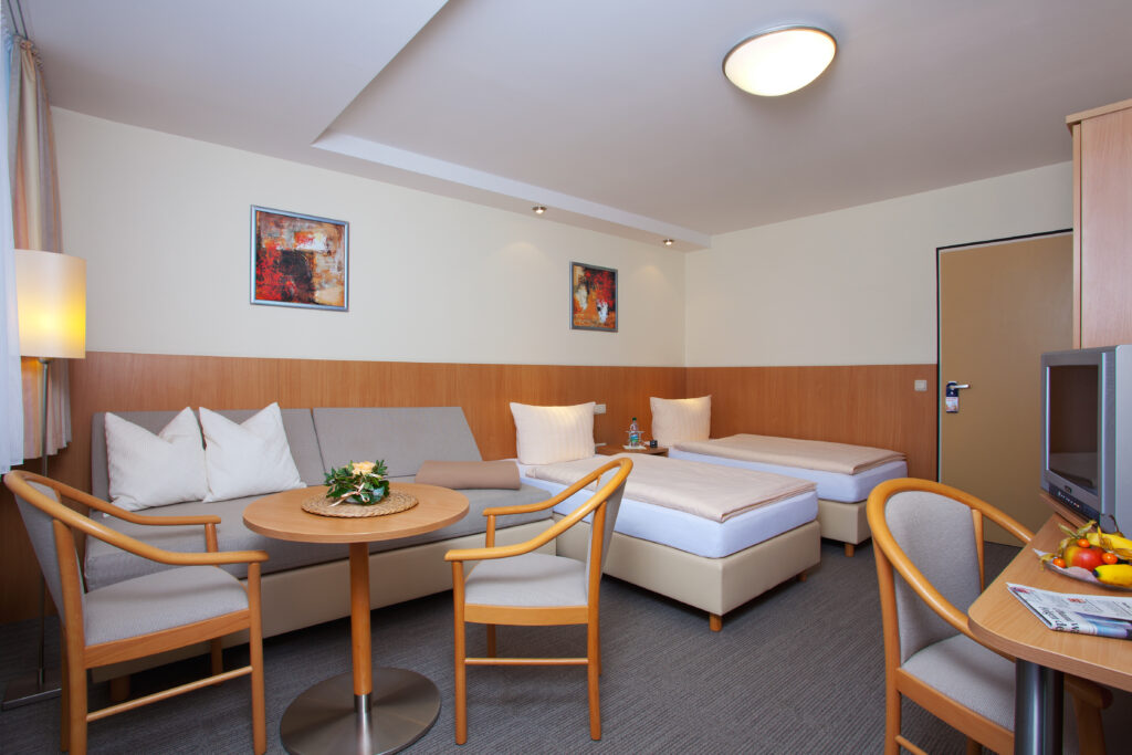 Hotelzimmer im Haus Silberbach mit Wohnzimmerbereich, Sofa, zwei Einzelbetten, Tisch und Stühle, Fernseher, Pflanzen und neutrale Beleuchtung und Holzboden