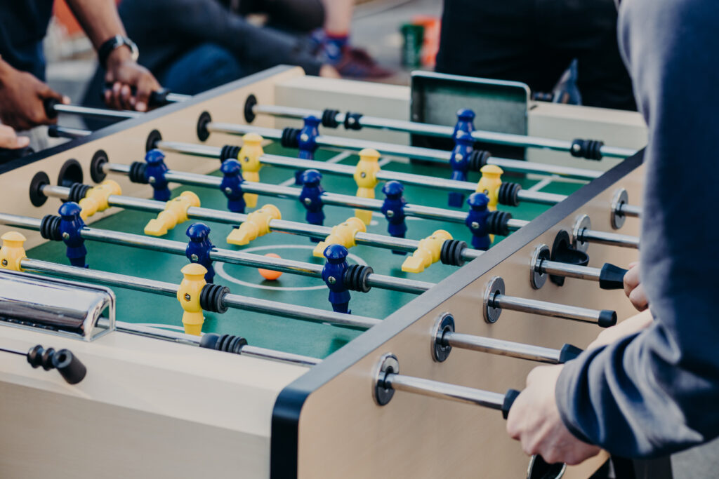 Foosball-Tisch im Freien mit grünem Spielfeld, blauen und gelben Spielsteinen, mehrere Spieler repetitiv am Spiel beteiligt; Kontext: Gruppenaktivität bei Haus Silberbach im Fichtelgebirge in Oberfranken