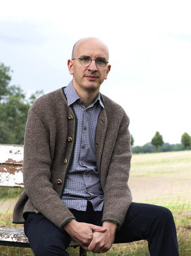 Porträt von Hein Jakob in brauner Strickjacke bei der Kulturreihe im Hotel Morgenland vor einer ländlichen Landschaft